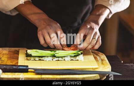 Hände kochen, japanische Sushi-Rolle machen. Der Prozess der Zubereitung von Sushi-Rollen mit Lachs, Gurke, Reis und Nori. Stockfoto