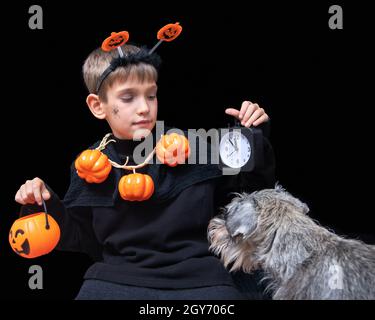 Halloween Lifestyle. Ein Junge mit einer Spinne auf der Wange und einer Kürbisperlen mit einem orangefarbenen Halloween-Korb mit Pralinen und einem schwarzen Wecker A Stockfoto