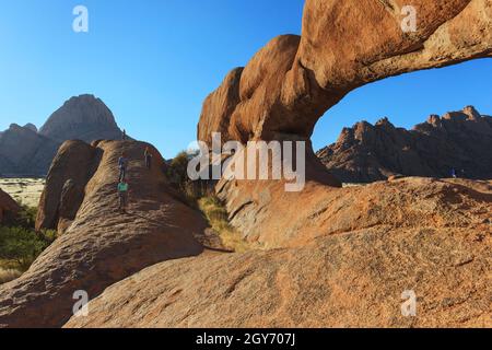 Reisende, Touristen erklimmen die Rock Bridge, den natürlichen Bogen des Spitzkoppe-Viertels. Wunderschöner blauer Himmel und warme Farben. Damaraland, Namibia, Afrika Stockfoto