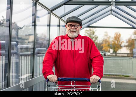 Lächelnder Senior mit einer Mütze und einer roten Jacke, die einen Einkaufswagen drückt Stockfoto