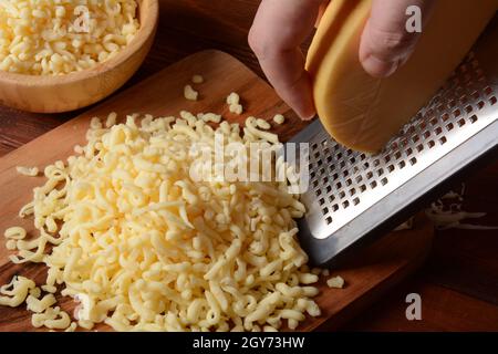 Mann, der Käse auf ein kleines Holzbrett geriebenhat. Geriebener Käse zum Kochen auf einem Schneidebrett auf einem hölzernen Hintergrund Stockfoto