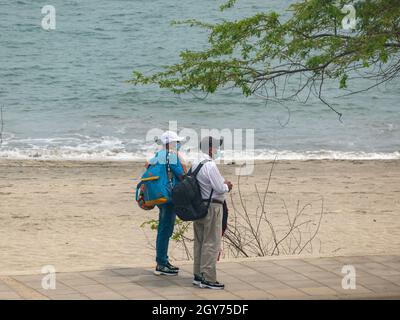 Santa Marta, Magdalena Kolumbien - 1. Juni 2021: Eine Frau und ein Mann stehen vor dem Meer mit Maske ihre Taschen in der Nähe des Flughafens Stockfoto