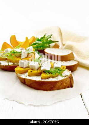 Bruschetta mit gebackenem Kürbis, gesalzenem Feta-Käse, Ricotta, Rucola und Gewürzen auf Pergament, Handtuch- und Gemüsescheiben auf Holzbrett-Hintergrund Stockfoto