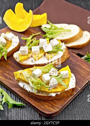 Bruschetta mit gebackenem Kürbis, gesalzenem Feta-Käse, Ricotta, Rucola und Gewürzen, Serviette und Gemüsescheiben auf Holzbrett Hintergrund Stockfoto