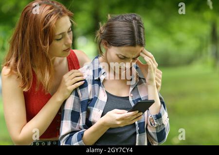 Ein trauriger Student, der auf dem Smartphone nachgeht und von einem Freund in einem Park oder auf dem Campus getröstet wird Stockfoto