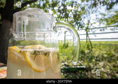 Hausgemachte Limonade aus Zitronen in einem großen Glaskrug auf dem Tisch im Garten. Ein Krug mit Zitrone und Minze steht auf der Straße gegen das Backdro Stockfoto