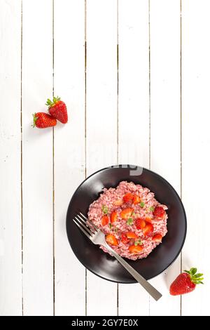 Risotto mit Erdbeerfrüchten serviert in einer schwarzen Schale auf weißem Holztisch. Flach liegend mit Kopierraum. Italienische Gourmetküche. Selektiver Fokus. Stockfoto