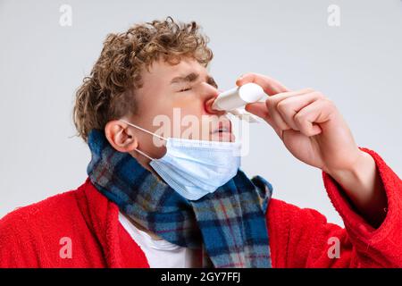 Nahaufnahme eines jungen Mannes mit Erkältung und Grippe, tropfende Nase mit Nasenspray isoliert auf grauem Hintergrund. Stockfoto