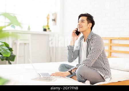 Hübscher junger Mann in legerer Kleidung, der zu Hause arbeitet, während er auf dem Bett sitzt, mit einem Laptop und im Schlafzimmer telefoniert. Stockfoto