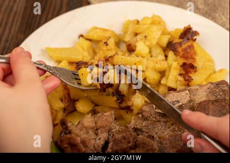 Jemand schneidet mit einem Messer und einer Gabel ein Stück saftiges Braten auf einem Teller gebratener Kartoffeln. Nahaufnahme Selektiver Fokus Stockfoto