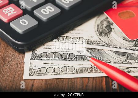 Dollar in 100-Dollar-Scheinen sind auf dem Tisch neben einem Rechner, einer Kreditkarte und einem Stift verteilt. Einhundert-Dollar-Scheine. US-Dollar. Stockfoto