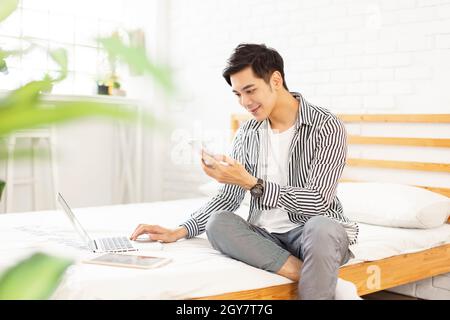 Hübscher junger Mann, der zu Hause arbeitet, während er mit einem Laptop und einem Smartphone auf dem Bett sitzt Stockfoto