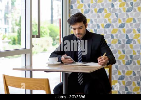 Kaukasischer Geschäftsmann in schwarzem Anzug, der während der Mittagspause eine Notiz auf dem Mobiltelefon liest. Atmosphäre in einem modernen Café. Stockfoto