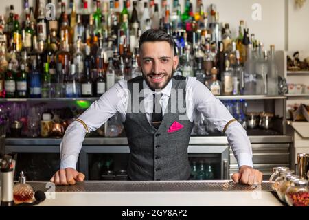 Eleganter junger bärtiger hispanischer Barkeeper in stilvoller Kleidung, lächelnd und mit Blick auf die Kamera, während er an der Theke in der modernen Bar mit Flaschen Alkohol arbeitete Stockfoto