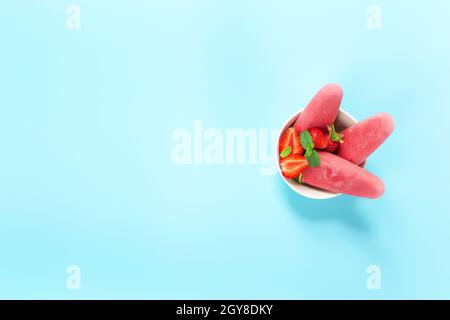 Draufsicht auf Schüssel mit fruchtigem, rosafarbenem, gefrorenem Sorbet-Eis auf Plastikstock mit Erdbeere, Minze auf blauem Hintergrund. Gefrorenes gemischtes Obst mit Joghurt i Stockfoto