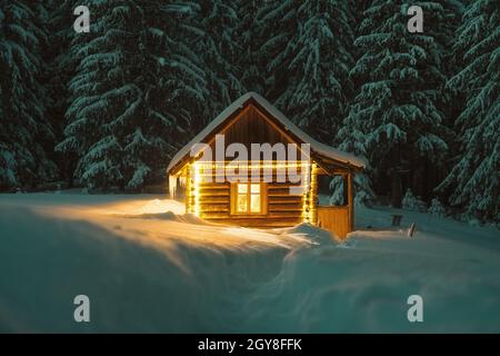 Fantastische Winterlandschaft mit glühender Holzhütte im verschneiten Wald. Gemütliches Haus in den Karpaten. Weihnachtsfeiertagskonzept Stockfoto