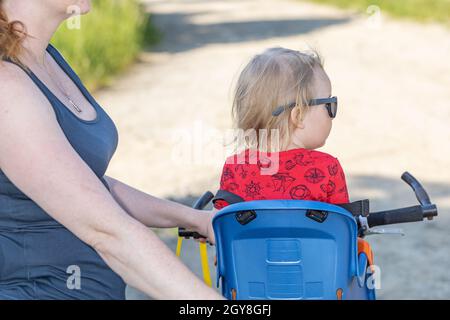 Rückansicht des kleinen Jungen, der auf einem Fahrradsitz ohne Schutzhelm sitzt. Seine Mutter hält Fahrrad. Stockfoto