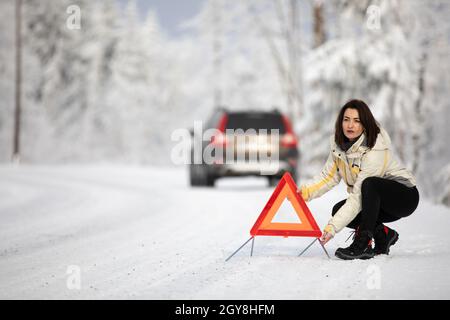 Eine hübsche, junge Frau, die ein Warndreieck aufstellte und um Hilfe rief, nachdem ihr Auto mitten im Nirgendwo bei einer eisigen Winterpause zusammengebrochen war Stockfoto