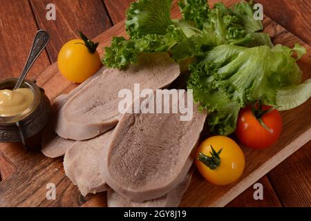 Geschnittene Rinderzungenscheiben auf einer Platte mit Salatblättern, Kirschtomaten und Dijon-Senf auf Holzgrund Stockfoto