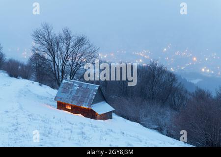 Fantastische Winterlandschaft mit glühendem Holzhaus vor dem Hintergrund glühender Lichter der Stadt im Nebel. Gemütliche Hütte in den Karpaten. Weihnachtsfeiertagskonzept Stockfoto