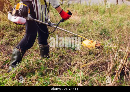 Gärtner mäht Unkraut mit dem Bürstenschneider auf der Herbstwiese. Arbeiter trimmen trockenes Gras mit manuellem Benzin-Trimmer mit Metallklinge Scheibe. Landwirt trägt b Stockfoto