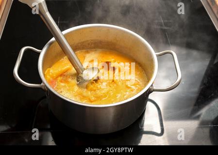 Vegetarische Gemüsesuppe von Hokkaido oder Red kuri Squash wird mit einem Stabmixer in einem Stahltopf auf einem schwarzen Herd püriert, im Herbst kocht Cop Stockfoto