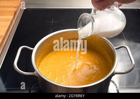 Kokosmilch oder Rahm in eine Gemüsesuppe aus rotem Kuri-Kürbis oder Hokkaido-Kürbis in einen Stahltopf auf einen schwarzen Herd gießen, ein Herbstgericht kochen, CO Stockfoto