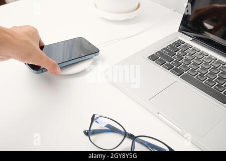 Frau, die Smartphone lädt Akku mit kabellosem Ladegerät in der Nähe von Laptop-Computer, moderne Technologie übertragen Energie Handy auf w Stockfoto