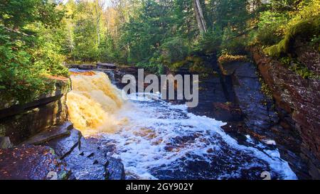 Blauer Fluss mit goldenem und braunem Wasserfall in einer kleinen Schlucht mit Wald auf beiden Seiten Stockfoto