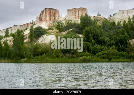 Wunderschöne Naturlandschaft. Weiße Kreidefelsen, Baumdickichte, ein wolkig bewölkter Himmel, ein ruhiger See am Fuße des Berges. Kalksteinbruch. Berg. Stockfoto