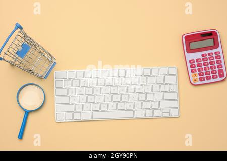 Weiße kabellose Tastatur und leerer Warenkorb, Lupe auf beigem Hintergrund, Budget-Analyse-Konzept, Einsparungen. Online-Shopping Stockfoto