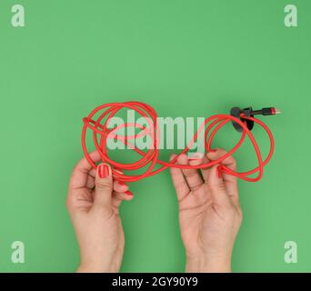 Weibliche Hand hält verdrilltes Kabel zum Laden von mobilen Geräten in einer roten Textilverpackung auf grünem Hintergrund, Nahaufnahme Stockfoto