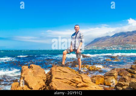 Männlicher Reisevorführer in False Bay raue Küstenlandschaft mit Felsbrocken Wellen und Bergen mit Wolken in Glencairn Simons Town Kapstadt Wester Stockfoto