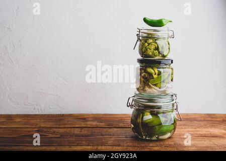 Drei Glas Jalapeno Peppers aus der frischen Dose mit Kräutern und Knoblauch auf einem Holztisch. Speicherplatz Kopieren Stockfoto