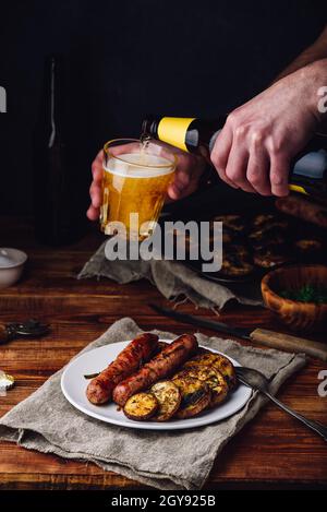 Schweinswürste gebacken mit Auberginen, Lauch und Kräutern auf weißem Teller. Mann gießt Bier aus der Flasche in ein Glas. Stockfoto