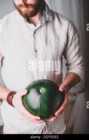 Bärtiger Mann im Leinenhemd hält in seinen Händen ganz grüne Wassermelone Stockfoto
