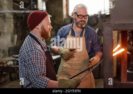 Schmied in Handschuhen schmiedet zusammen mit dem Meister Eisen während ihrer Arbeit in der Werkstatt Stockfoto