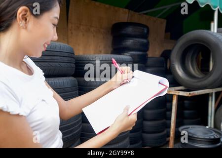 Roter Stift, Papier und Zwischenablage im Besitzer der Autoteile speichern Hand. Mehrere Autoreifen sind in einem Reifenladen aufgereiht. Stockfoto