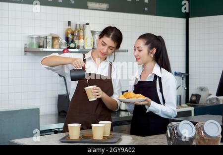 Der junge kaukasische Barista gießt Milch aus dem Krug in eine Papierkaffeebecher. Asiatische Assistentin in einer Schürze, die einen Croissantteller hält, steht neben ihr. Morn Stockfoto