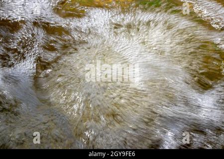 Wasser wirbelt in einem Bach, der über felsigen Boden fließt und von der Sonne reflektiert wird. Makro. Stockfoto