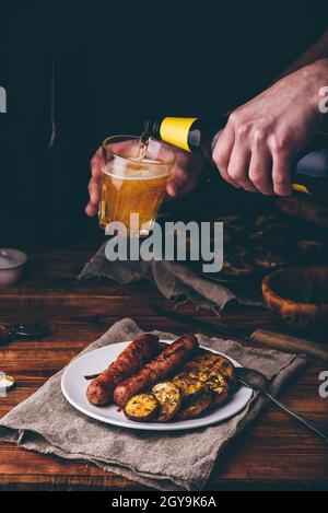 Schweinswürste gebacken mit Auberginen, Lauch und Kräutern auf weißem Teller. Mann gießt Bier aus der Flasche in ein Glas. Stockfoto