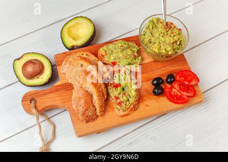 Frisch zubereitete Guacamole in kleiner Glasschüssel, Brot, Tomaten, Oliven am Arbeitsbrett und zwei Avocados-Hälften weiter, Blick von oben. Stockfoto