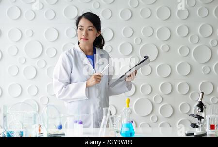 Junger asiatischer Wissenschaftler mit Stift und Zwischenablage. Sie schaute nach draußen und dachte an etwas. Arbeitsatmosphäre im chemischen Labor. Stockfoto