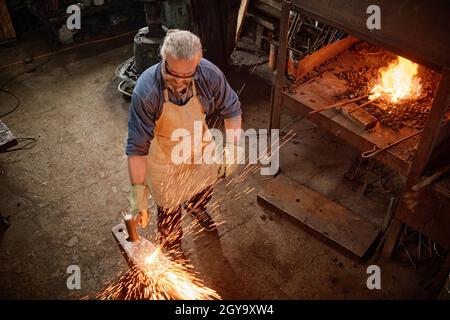 Hochwinkelansicht eines reifen Mannes in der Schürze, der das Metall in der Schmiede schmiedet Stockfoto