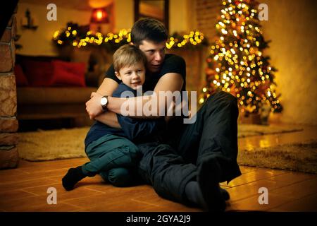 Der junge Vater umarmt seinen Sohn in einem Raum, der mit einem festlichen weihnachtsbaum und Girlanden am Silvesterabend geschmückt ist. Selektiver Weichfokus, Filmkorneffekt Stockfoto