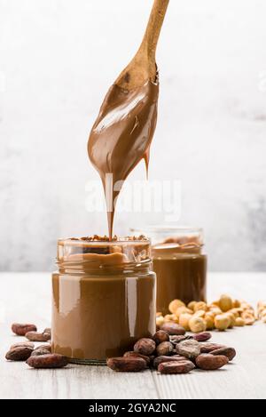 Schokoladencreme im Glas mit Kakaobohnen, Haselnüssen und tropfendem Löffel auf Holztisch Stockfoto