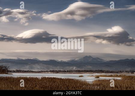 An einem kalten Wintertag erhebt sich ein Berg (Longs Peak) über einem gefrorenen See. Einzigartige Wolken hängen über den Bergen, während Schilf den See umschlang. Stockfoto