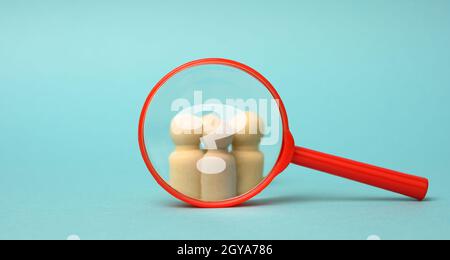 Holzfiguren von Männern und eine rote Lupe auf blauem Hintergrund. Rekrutierung für das Unternehmen, Gleichgesinnte und Teamarbeit. Suchen Sie nach Märchen Stockfoto