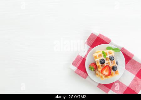 Weißer Teller auf rotem Küchentuch mit hausgemachten belgischen Waffeln, Heidelbeere, Erdbeerschnitt, Minzblatt, Zuckerpulver auf grauem Hintergrund Stockfoto