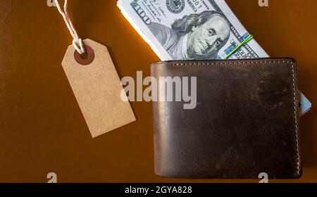 Viele Hunderte von Dollar-Banknoten in einer Lederbörse mit einem braunen Anhänger, Preisschild auf einem braunen Hintergrund. Flaches Lay. Hintergrund, Urlaubskonzept. Schwarz Stockfoto